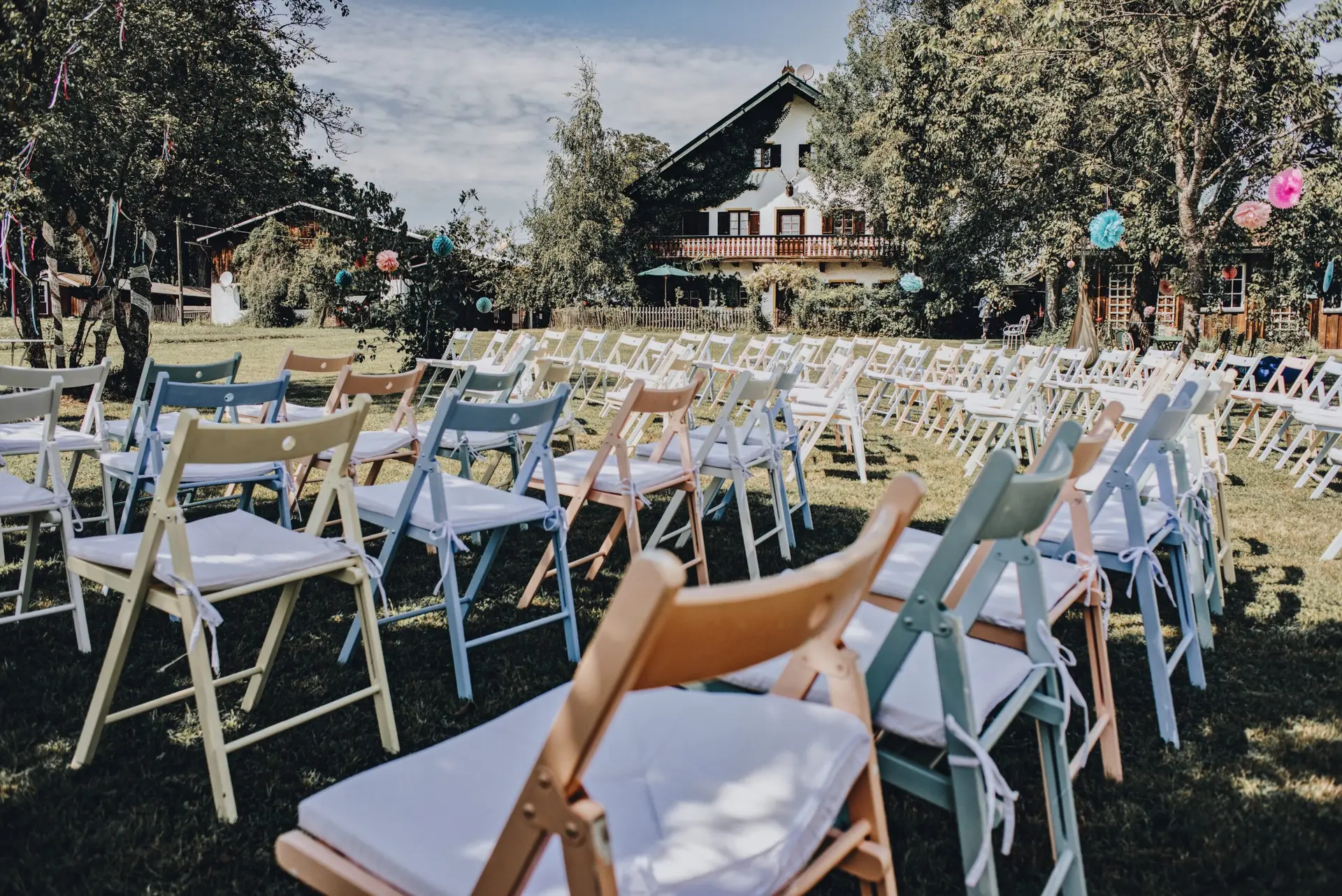 Sitzplätzchen in Bunt mieten Pastellfarbene Holzklappstühle in Weiß, Blau, Rosa und Natur in Reihen vor einem Landhaus mit Lampions